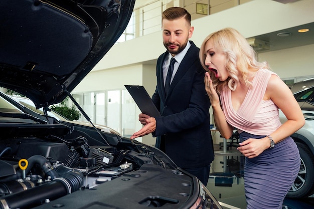 Caucasian salesman in suit showing catalog to beautiful female blonde welldressed customer in dealership salon