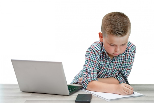 Caucasian school-age boy in a plaid shirt sitting at the table and writing in a piece of paper