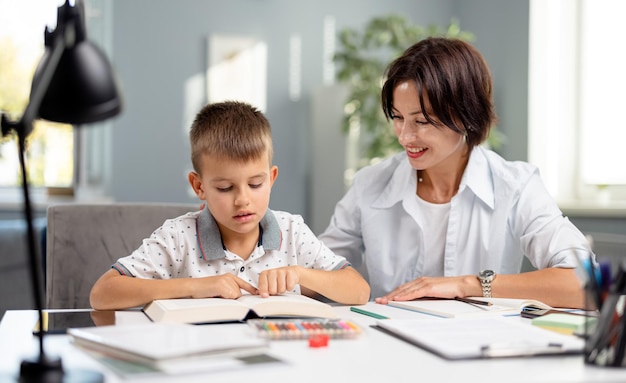 Caucasian schoolboy sitting with mother at desk and reading book Caring woman helping son with homework Educational moments during parenting