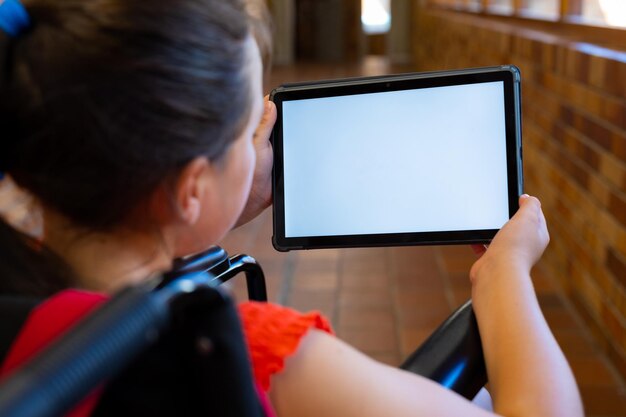 Photo caucasian schoolgirl sitting in wheelchair and using tablet with copy space at school corridor