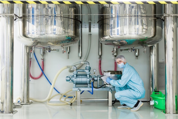 Caucasian scientist in blue lab suit sit under big steel barrels work with blue engine look at camera