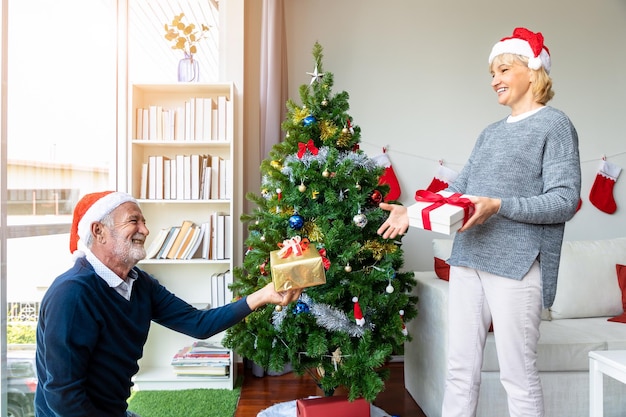 Caucasian senior elderly man hand gift box to woman while decorating Christmas tree together for the festival