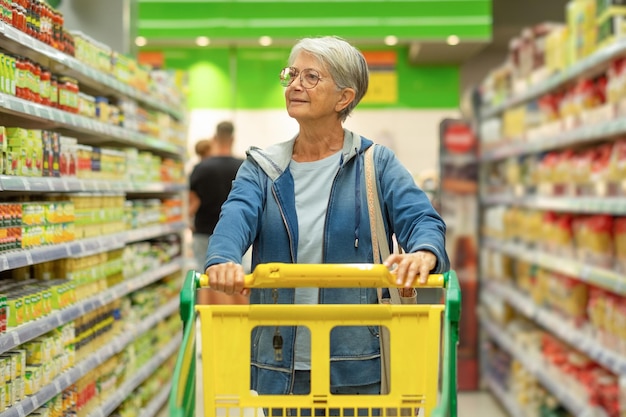 ディスプレイ上の製品を見てスーパーマーケットでトロリーを押す白人の年配の女性消費者購入コンセプト