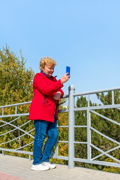 Donna maggiore caucasica in giacca rossa utilizza lo smartphone nel parco