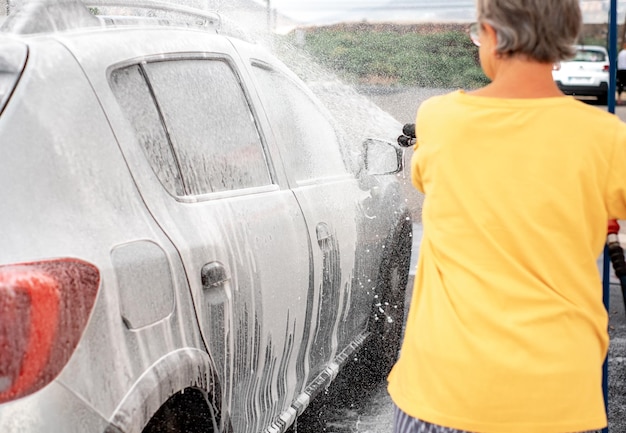 Donna anziana caucasica che lava la sua auto in una stazione di lavaggio auto self-service utilizzando acqua ad alta pressione autolavaggio self-service