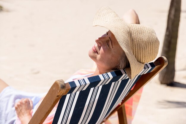 사진 모자를 쓰고 눈을 감고 해변의 갑판 의자에서 자고 있는 백인 고위 여성. 공간, 변경되지 않은, 은퇴, 휴가, 생활 방식, 즐거움, 휴식 및 여름 개념을 복사합니다.