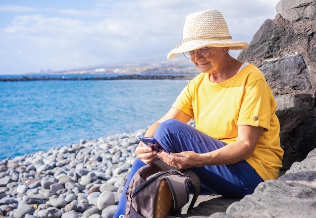 휴대 전화를 사용하여 바다의 자갈 해변에 앉아 밀짚 모자를 쓰고 백인 수석 여성 기술 및 사회적 개념을 사용하여 휴대폰을 들고 성숙한 여성 휴가 자유와 행복한 은퇴의 개념