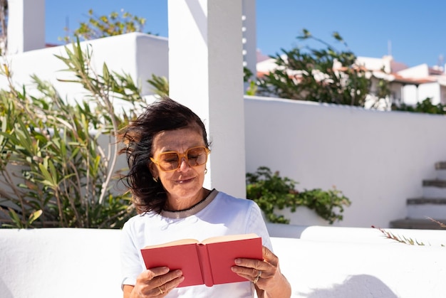 Caucasian senior woman with sunglasses relaxing sitting in a bench with a book enjoying a sunny day