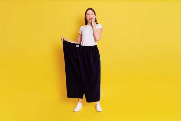 Caucasian shocked female with dark hair looking at camera with big eyes and open mouth, covering cheek with palm, wearing too big pants, woman lost weight, isolated over yellow wall.