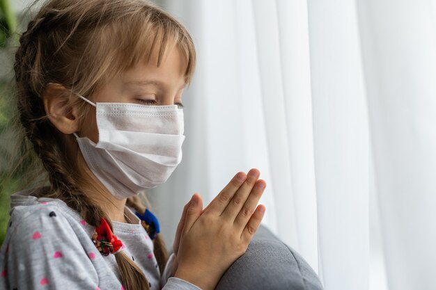 Caucasian sick little girl in medical mask during coronavirus epidemic prays