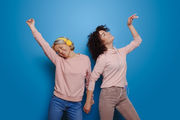 Caucasian sisters full of emotions listening to music using headphones on a blue  wall