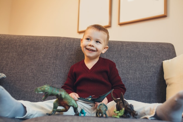 Caucasian small boy sitting on the sofa and playing with his\
dinosaur toys while looking at his parents