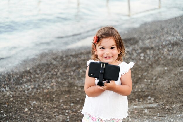 Caucasian small girl with selfie stick on the seaside. Taking photo, recording vlog, video call