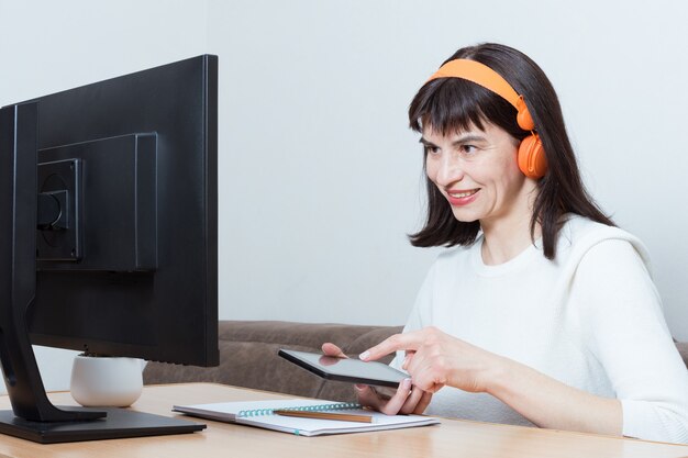 Donna sorridente caucasica con cuffie arancioni seduta davanti al monitor e con in mano uno smartphone per lo shopping, la ricerca di informazioni, la formazione online