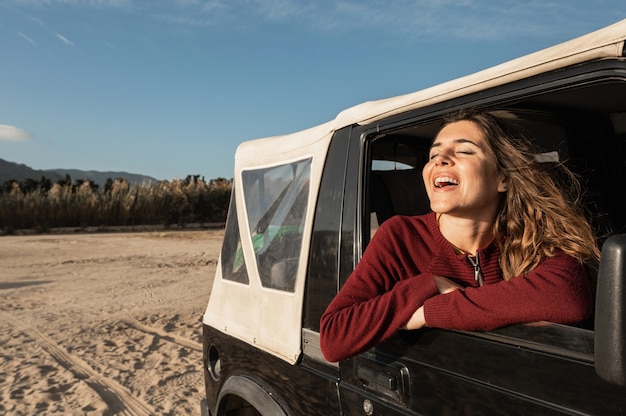 Caucasian smiling young woman with closed eyes looking out the window. Journey in Off-road 4x4 vehicle. Background of sandy desert and beach.