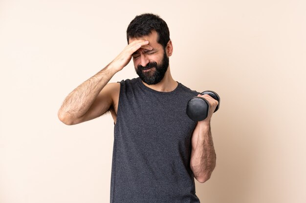 Uomo caucasico di sport con la barba che fa sollevamento pesi sopra la parete isolata con mal di testa