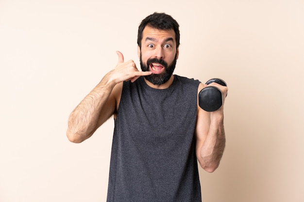 Caucasian sport man with beard making weightlifting over wall making phone gesture and doubting