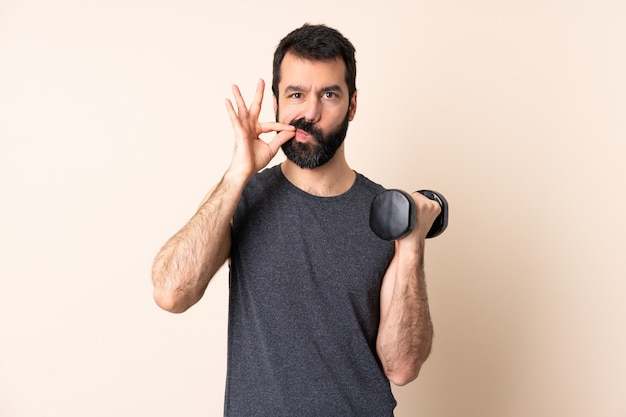 Uomo caucasico di sport con la barba che fa sollevamento pesi sopra la parete che mostra un segno di gesto di silenzio