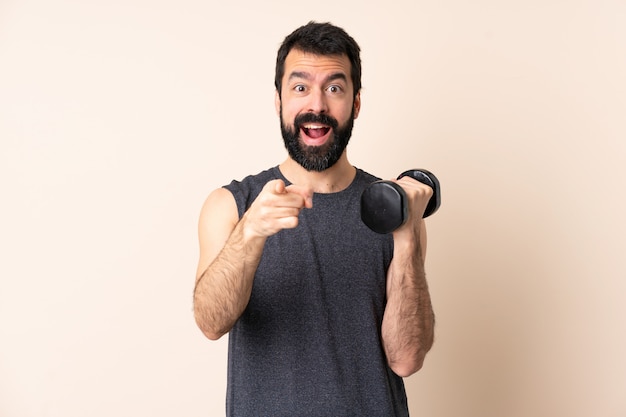 Uomo caucasico di sport con la barba che fa sollevamento pesi sopra la parete sorpresa e che indica parte anteriore