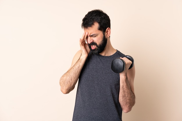 Uomo caucasico di sport con la barba che fa sollevamento pesi sopra la parete con mal di testa