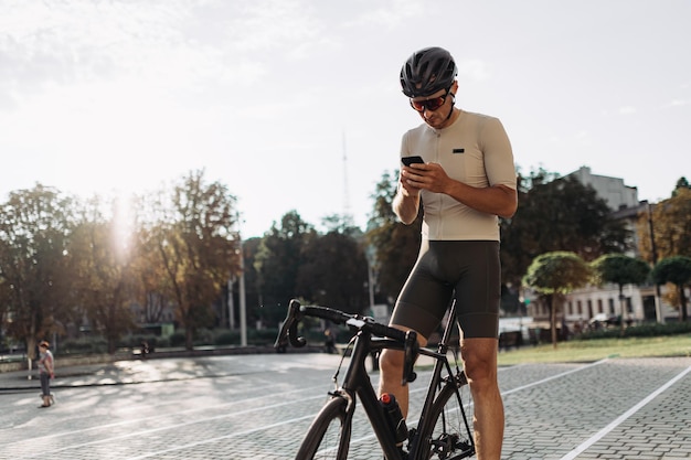 Caucasian sportsman in helmet and glasses using modern smartphone while resting after riding Male cyclist sitting on black bike among city street with gadget in hands