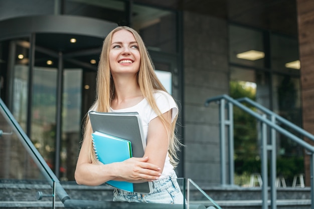 白人学生の女の子は、大学の建物の背景にフォルダー、ノートブックを手に持ち、笑顔で目をそらします。コピースペース