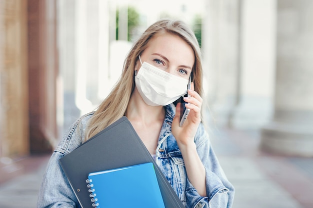 Caucasian student girl wearing a medical mask stands in front of the university and talks on a cell phone during coronavirus quarantine, covid-19