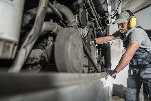 Photo caucasian technician in safety headphones thoroughly checking the condition of the engine in the bus during annual inspection automotive industry theme