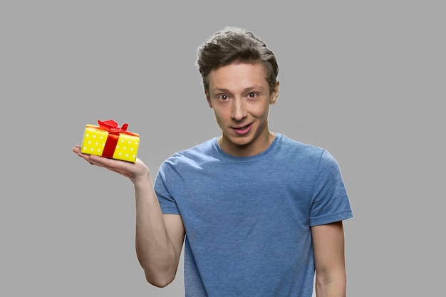 Caucasian teen boy holding small yellow gift box. Teenage guy holding present box on his palm against gray background.