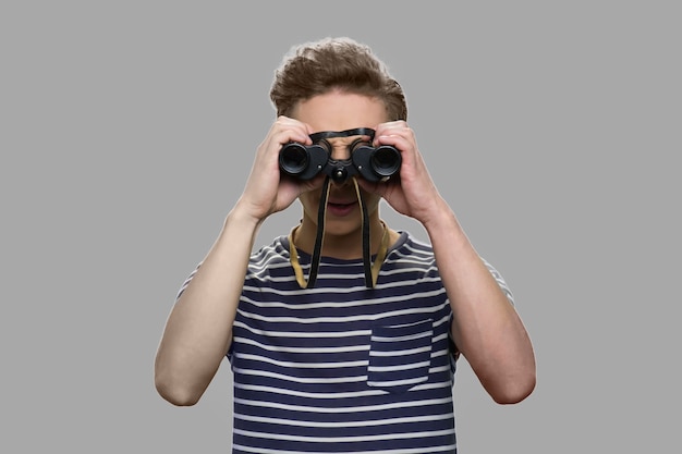 Caucasian teen boy looking through binoculars. Curious teen guy using binoculars standing against gray background.