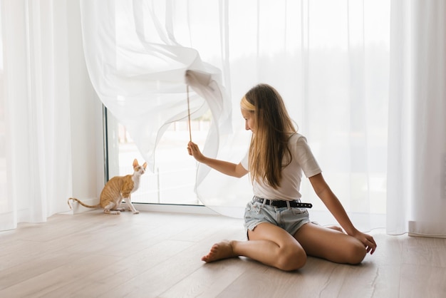 Caucasian teen girl sitting on the floor playing with a cat cornish rex with a stick teaser in the house