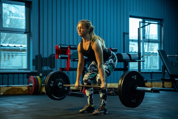 Photo caucasian teenage girl practicing in weightlifting in gym. female sportive model training with barbell, looks concentrated and confident. body building, healthy lifestyle, movement and action concept.