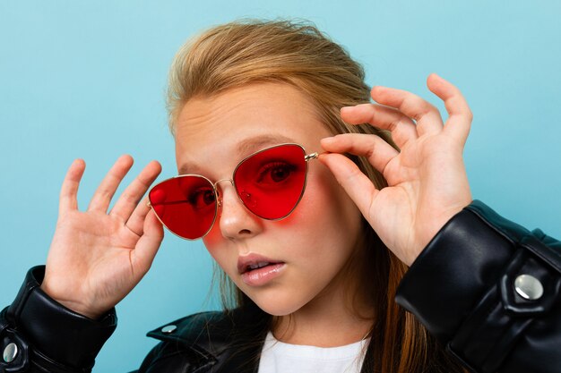 Caucasian teenager girl with brown hair in black jacket holds red sunglasses isolated on blue background