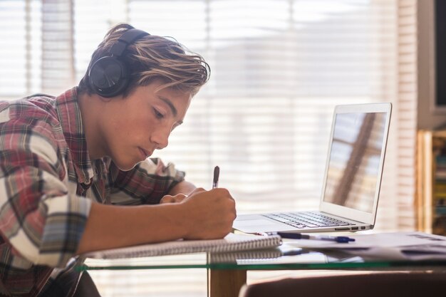 Caucasian teenager indoor doing homework on the table at home - blonde guy writing and reading in his laptop or computer to get greats scores