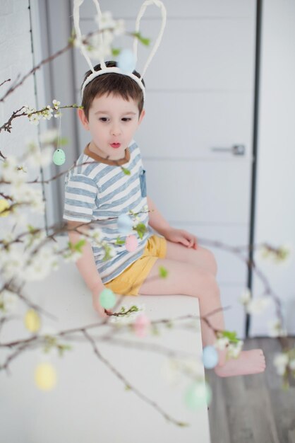 Caucasian toddler boy with funny bunny ears near easter tree with colorful eggs on a white background