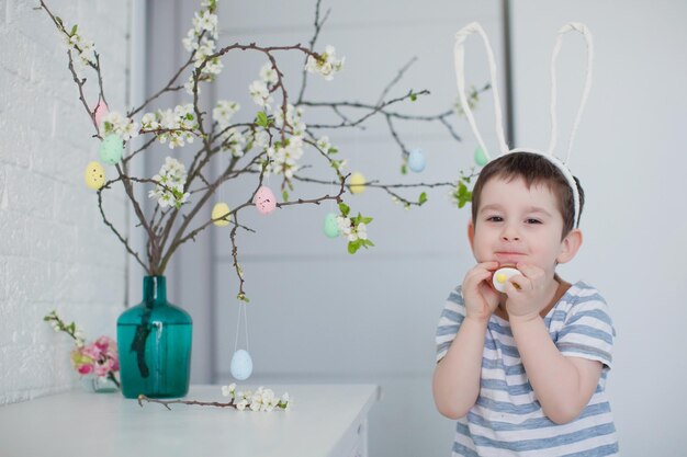 Caucasian toddler boy with funny bunny ears stands near easter tree with colorful eggs on a white background