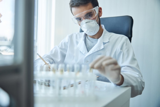 Caucasian virologist in safety goggles and a lab coat staring at the test tube rack