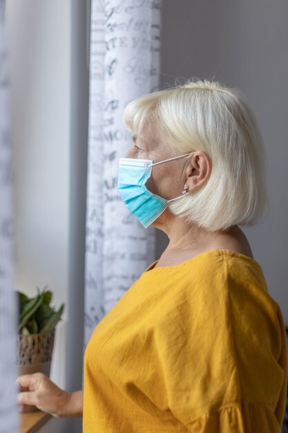 코로나 바이러스 전염병 동안 집이나 카린에 병원에 앉아있는 동안 창 밖을 바라 보는 보호 의료 마스크에 50 세 백인 여자.