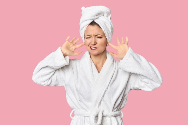 Белая женщина в халате и полотенце, закрывающая уши пальцами, напряженная и отчаянная.