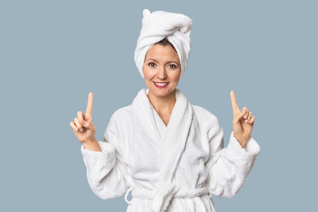 Caucasian woman in bathrobe and towel indicates with both fore fingers up showing a blank space