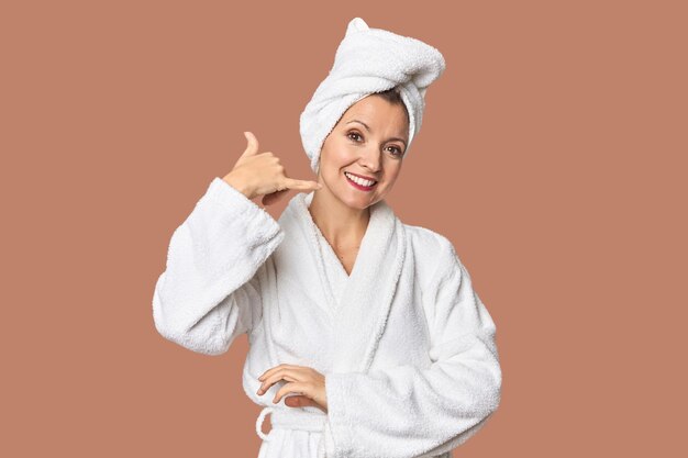 Caucasian woman in bathrobe and towel showing a mobile phone call gesture with fingers