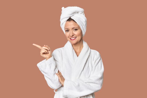 Photo caucasian woman in bathrobe and towel smiling cheerfully pointing with forefinger away