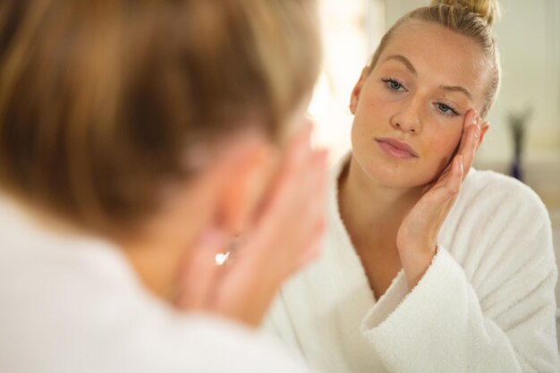 Caucasian woman in bathroom wearing bathrobe, looking in mirror and moisturising face. health, beauty and wellbeing, spending quality time at home.