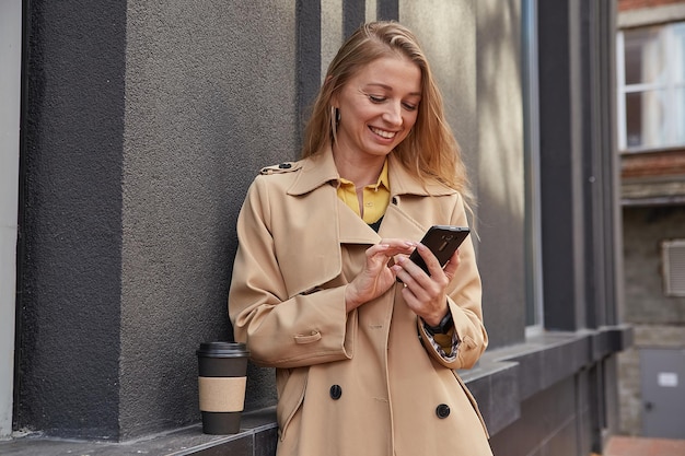 晴れた日に屋外でスマートフォンを使用してベージュのトレンチコートで白人女性