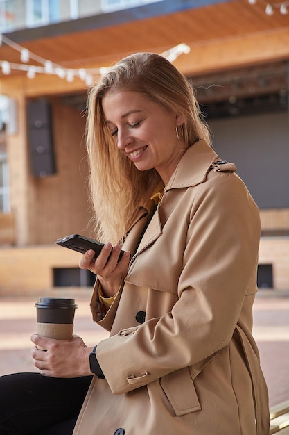 晴れた日に屋外でスマートフォンを使用してベージュのトレンチコートで白人女性