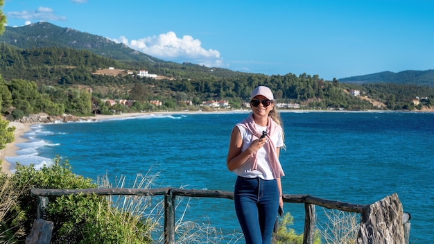 Кавказская женщина в кепке и солнцезащитных очках на смотровой площадке на побережье Эгейского моря Греции, скалистых холмах с растущими деревьями и кустами, зданиями