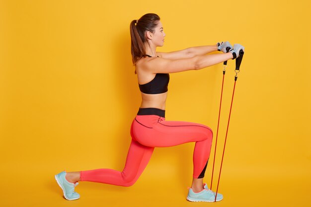 Caucasian woman coach in sporty short top and red gym leggings makes lunges by feet forward with fitness expander, female athlete stretch legs with elastic expander in pilates studio.