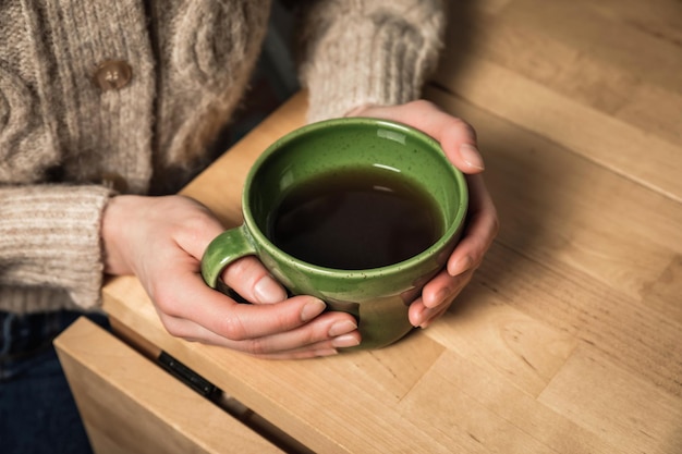 A caucasian woman in a cozy beige cardigan holding a green cup of tea in her hands sitting at the table a closeup image