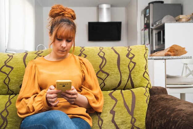 Photo caucasian woman dressed in yellow sitting on  sofa with the kitchen behind looking at her smartphone