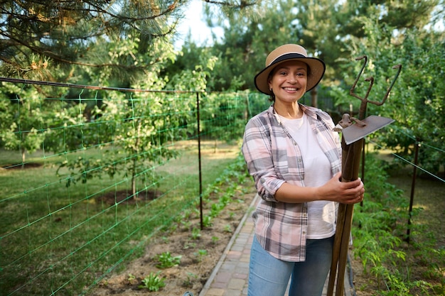 백인 여성 농부 원예가 정원사는 유기농 농장에서 정원 가꾸기 도구를 손에 들고 카메라를 향해 미소를 짓습니다.